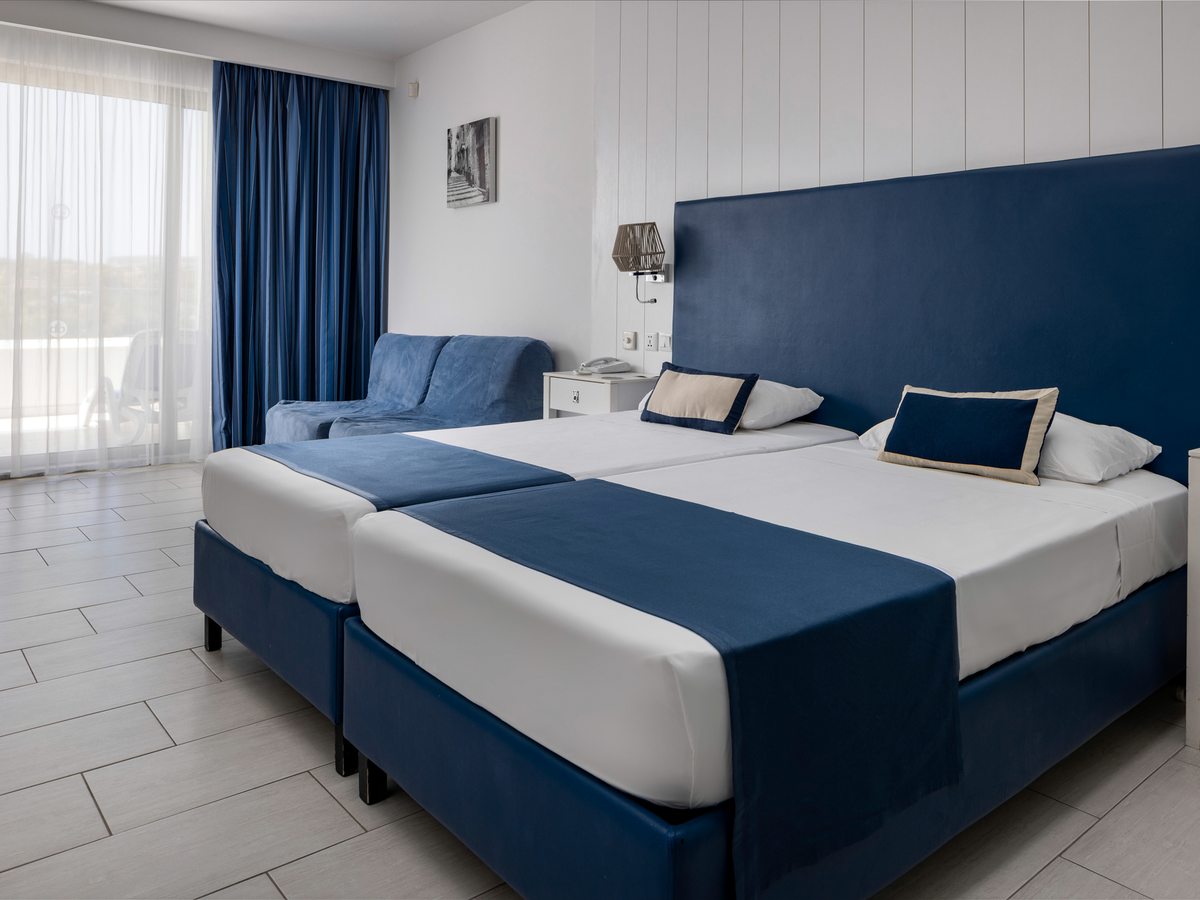 A room with two single beds pushed together under a blue headboard, a blue single sofa bed to one side, bedside lamps and a curtained balcony door.