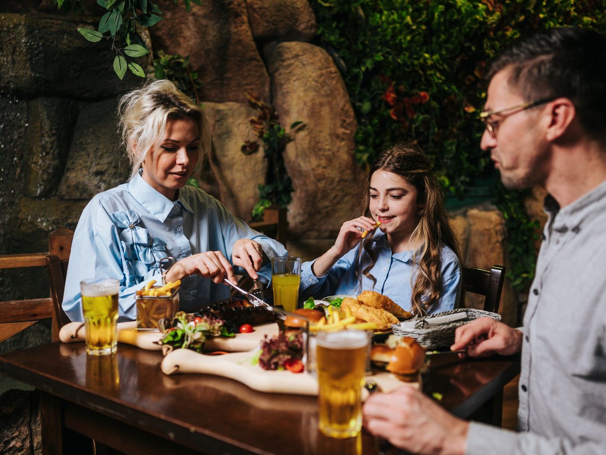 A jungle-themed restaurant with artificial tree trunks, rockwork and dense foliage around wooden tables, where a family sits over sharing plates and soft drinks.