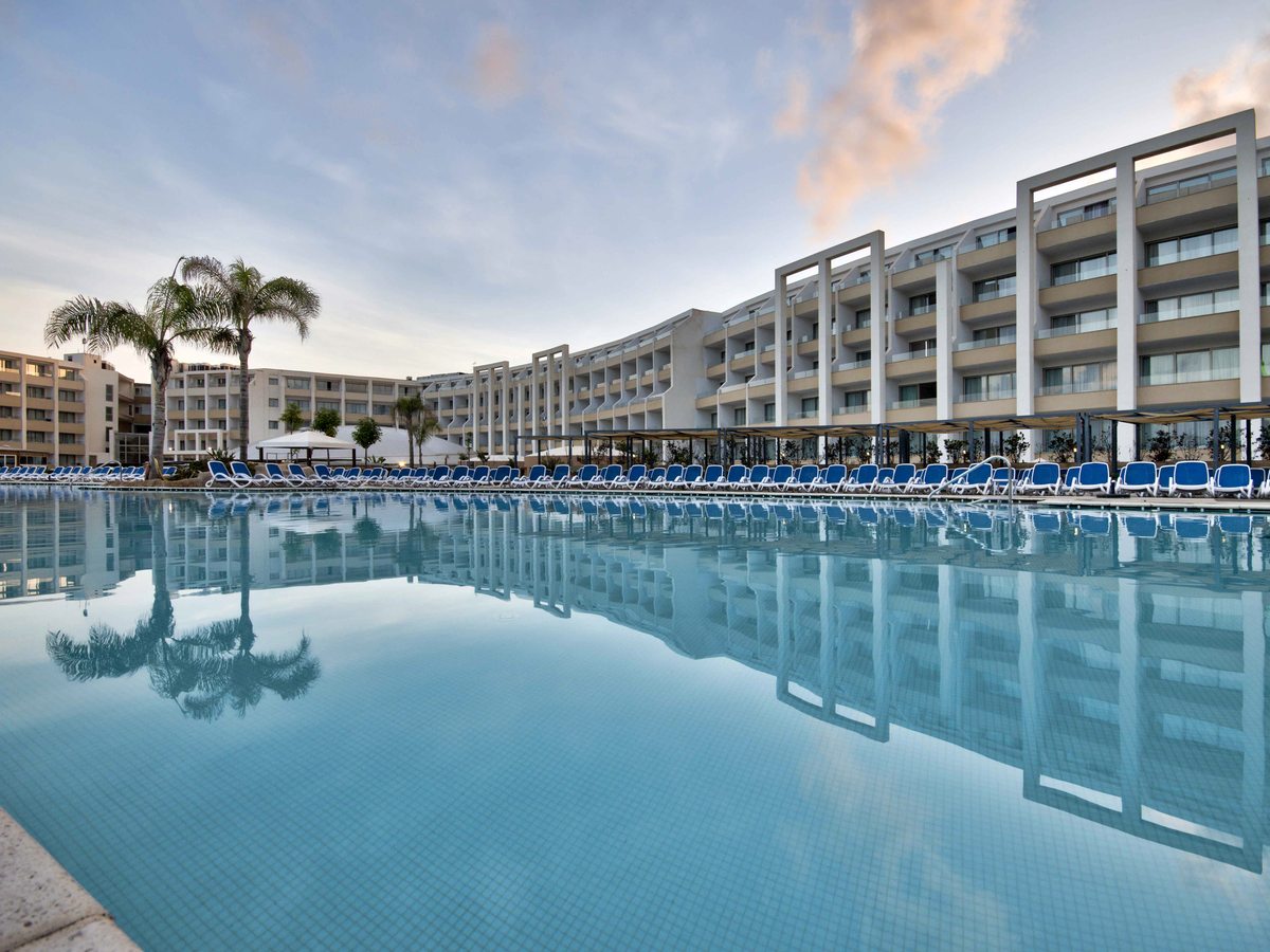 A large lagoon-style outdoor pool with rows of blue sun loungers along the tiled surround and the multi-storey resort behind under a soft sky.