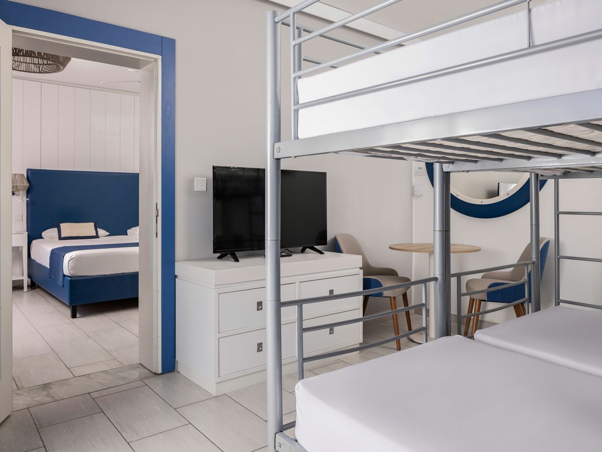 A family room with a metal bunk bed and, through an open doorway, a separate double bedroom with a television and a small dining table.