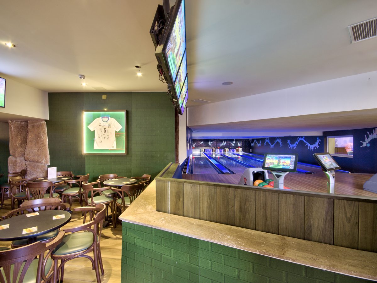 An indoor bowling alley of polished lanes with electronic scoring consoles, beside sports-bar seating with televisions and framed football shirts overhead.