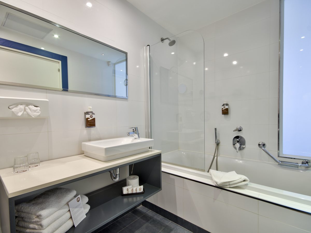 A bathroom with a bath and overhead shower behind a glass screen, a vessel basin on a wide grey vanity, a large mirror and folded towels.