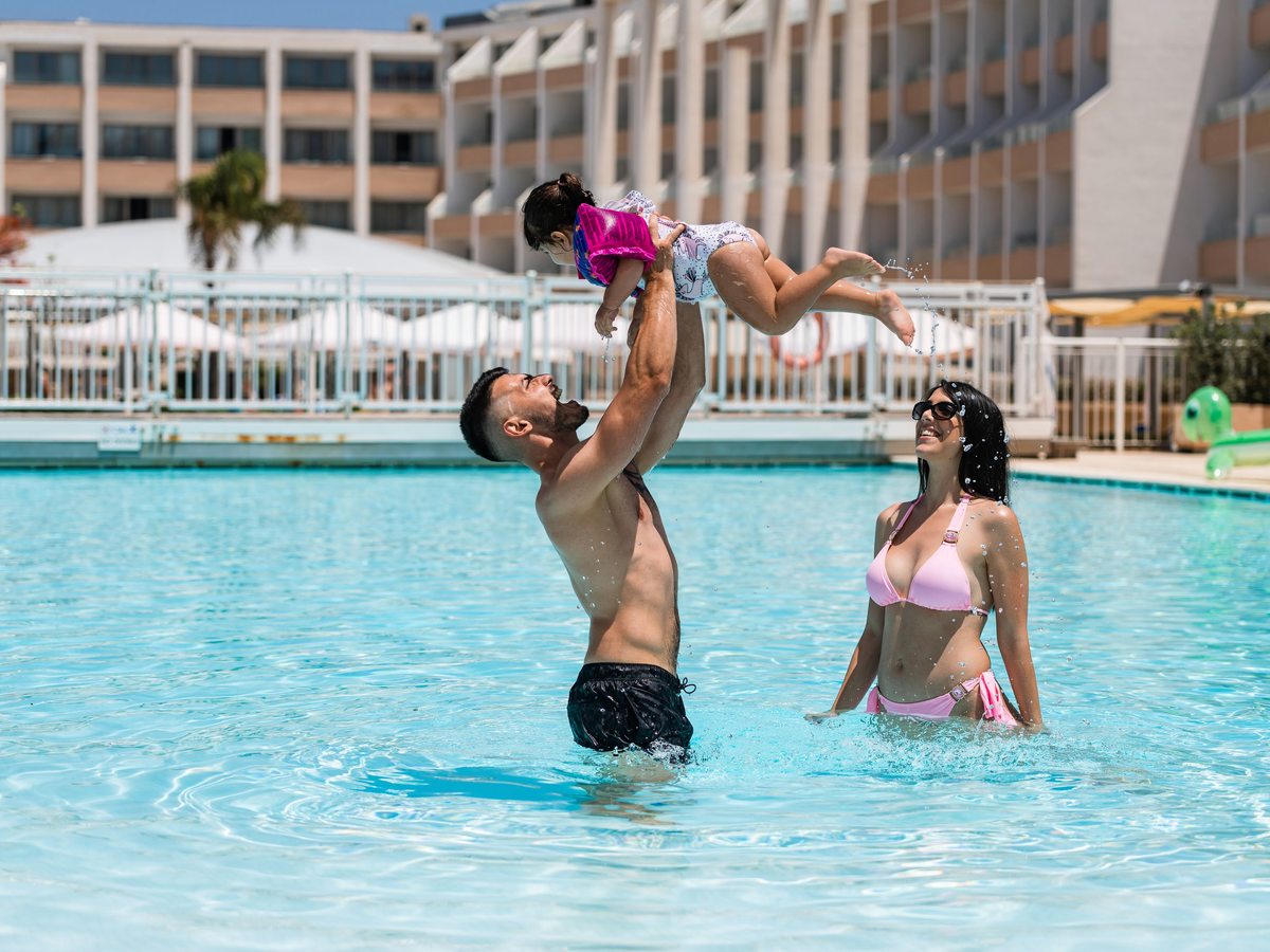 A father lifting a small child above the water in a bright outdoor pool, a woman beside them and the resort buildings behind.
