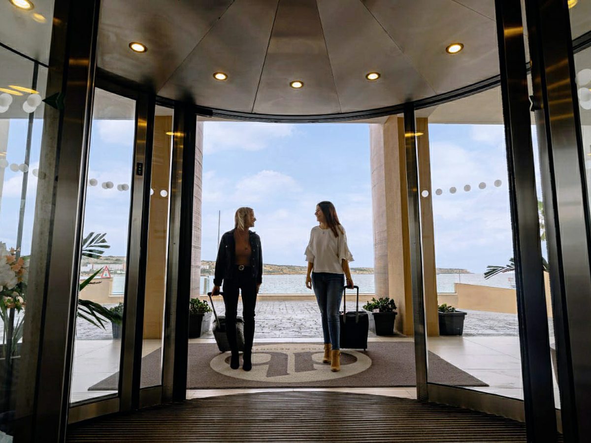 The resort's main entrance with a glass revolving door and a sea-facing terrace beyond, potted plants either side and two guests walking in across a tiled floor.