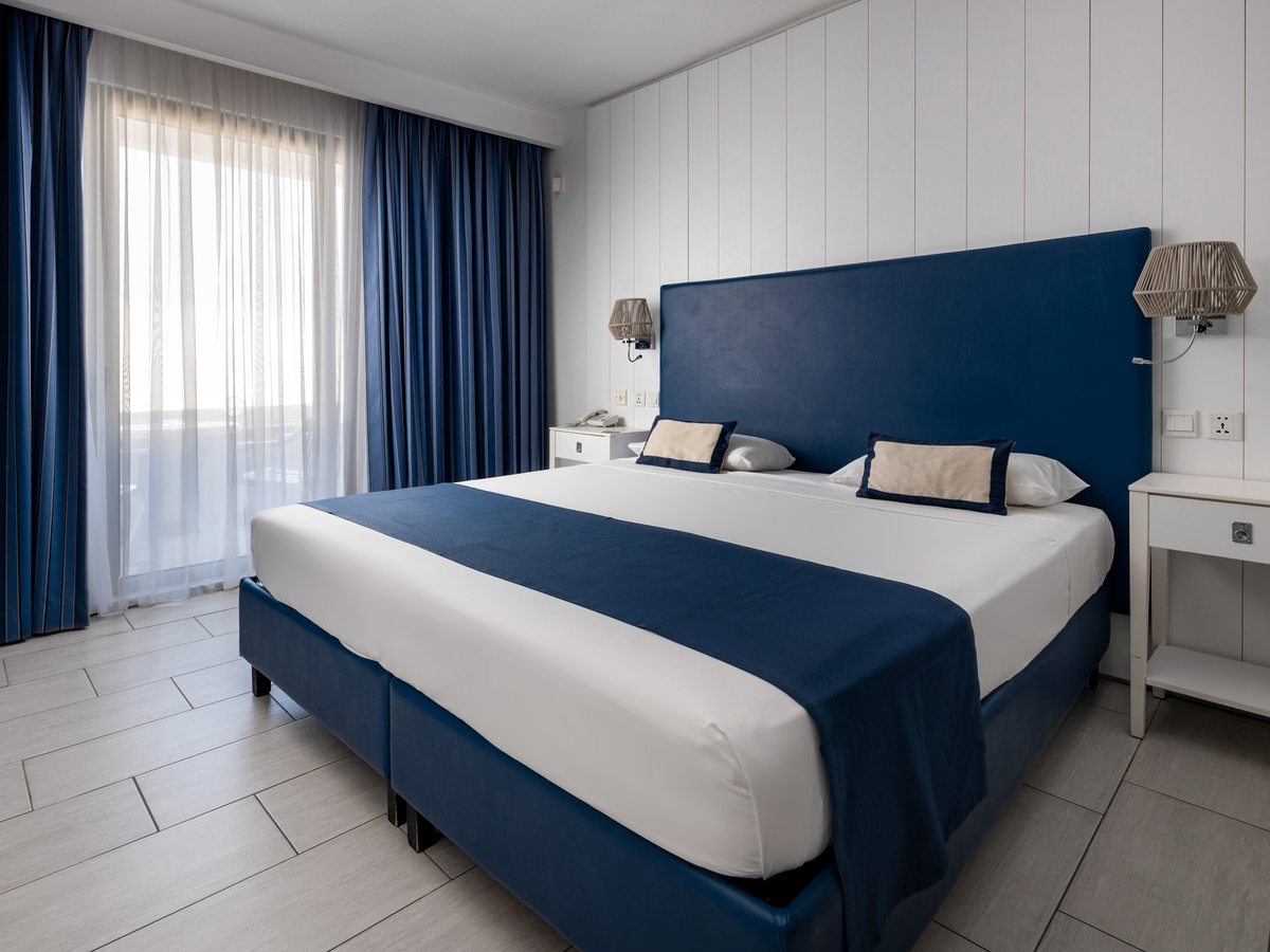 A guest room with a double bed against a tall blue upholstered headboard, bedside tables with reading lamps, blue curtains and a balcony door, on a tiled floor.