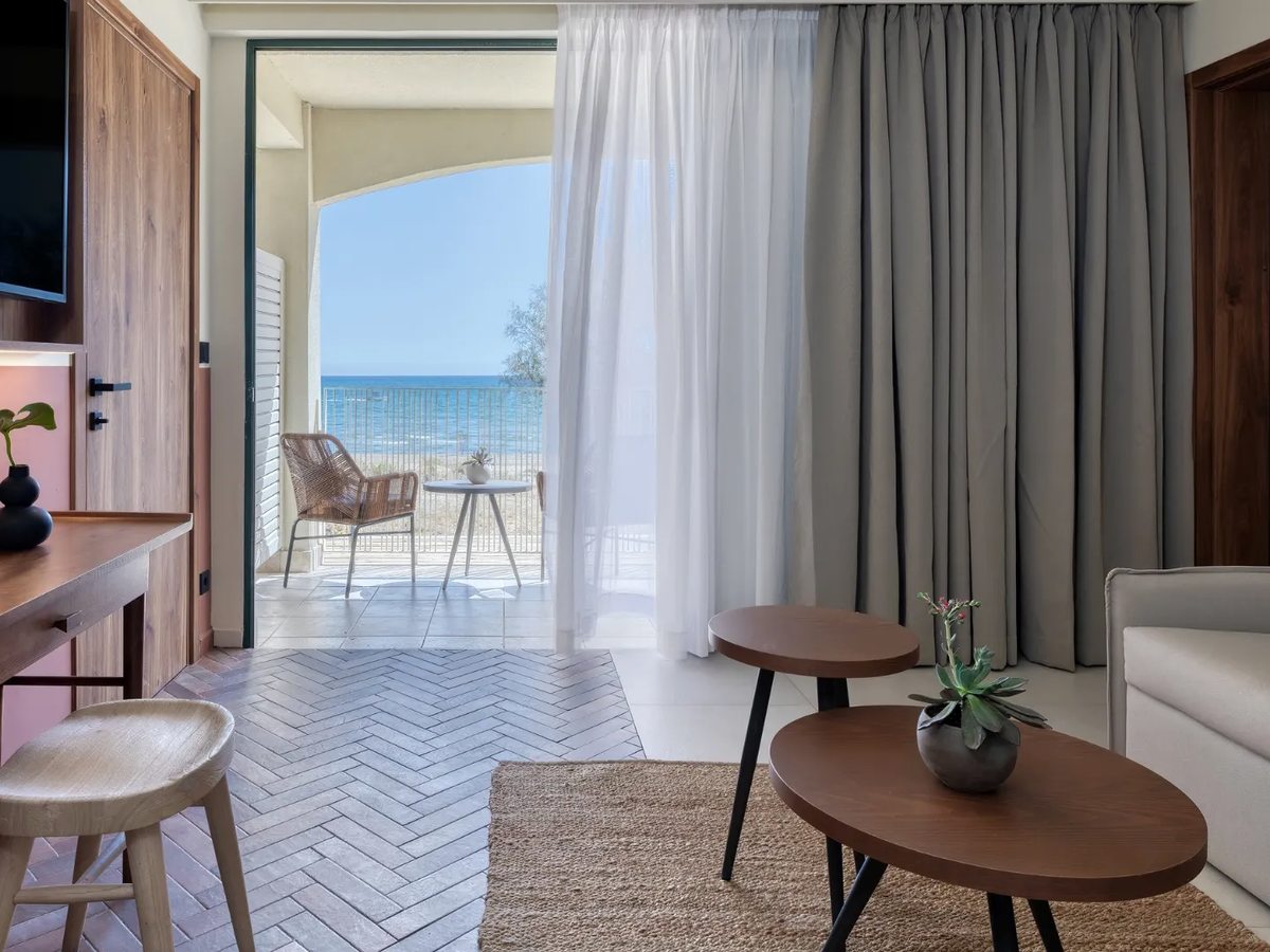 Suite living area with a sofa, round coffee tables and a desk beside sliding doors to a furnished sea-view balcony.