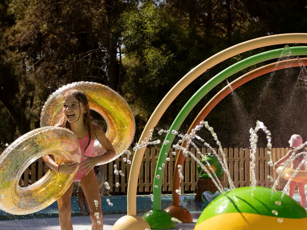 Children's splash park with rainbow spray arches and fountains over a shallow splash pad behind a low picket fence, with a child holding inflatable rings.