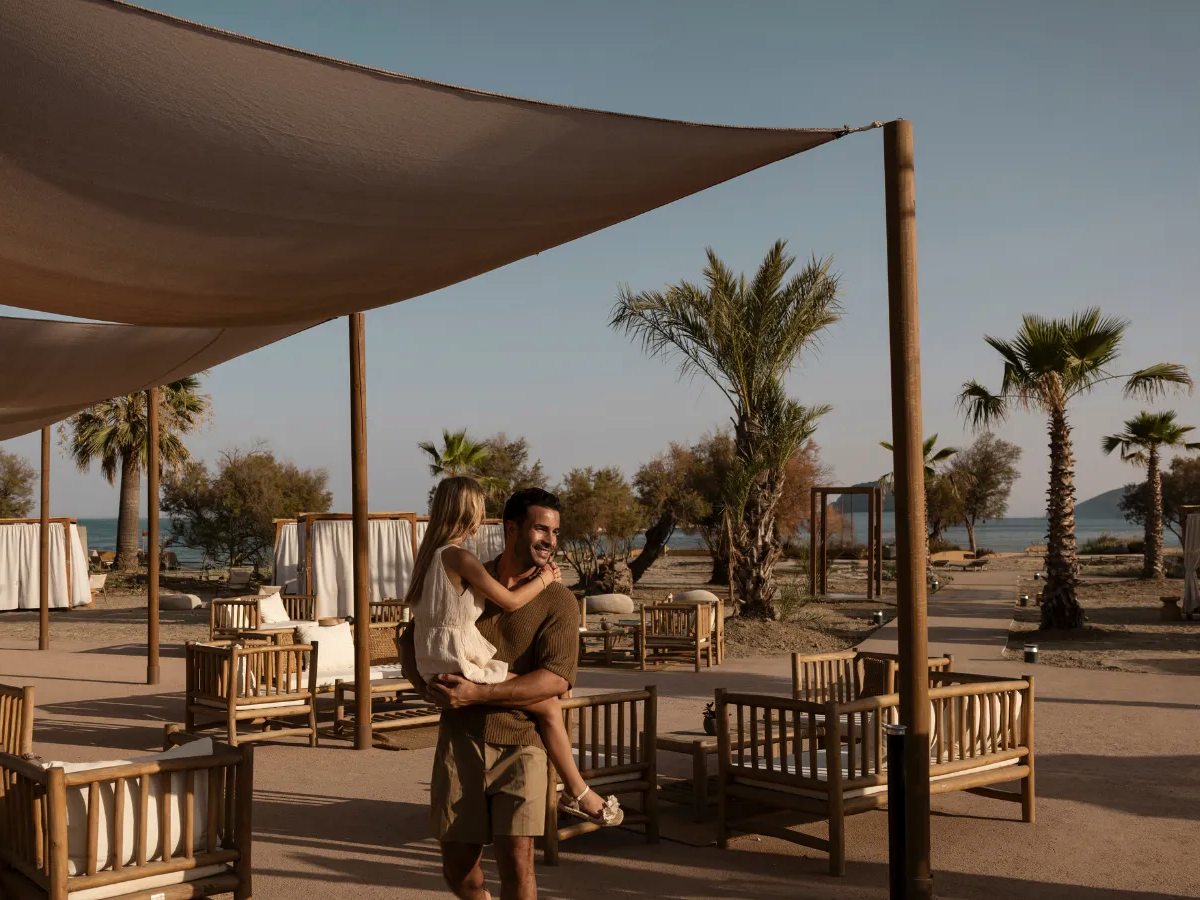 Beach lounge area with shade sails over timber benches, low tables and cabana beds among palm trees, with a glimpse of the sea.