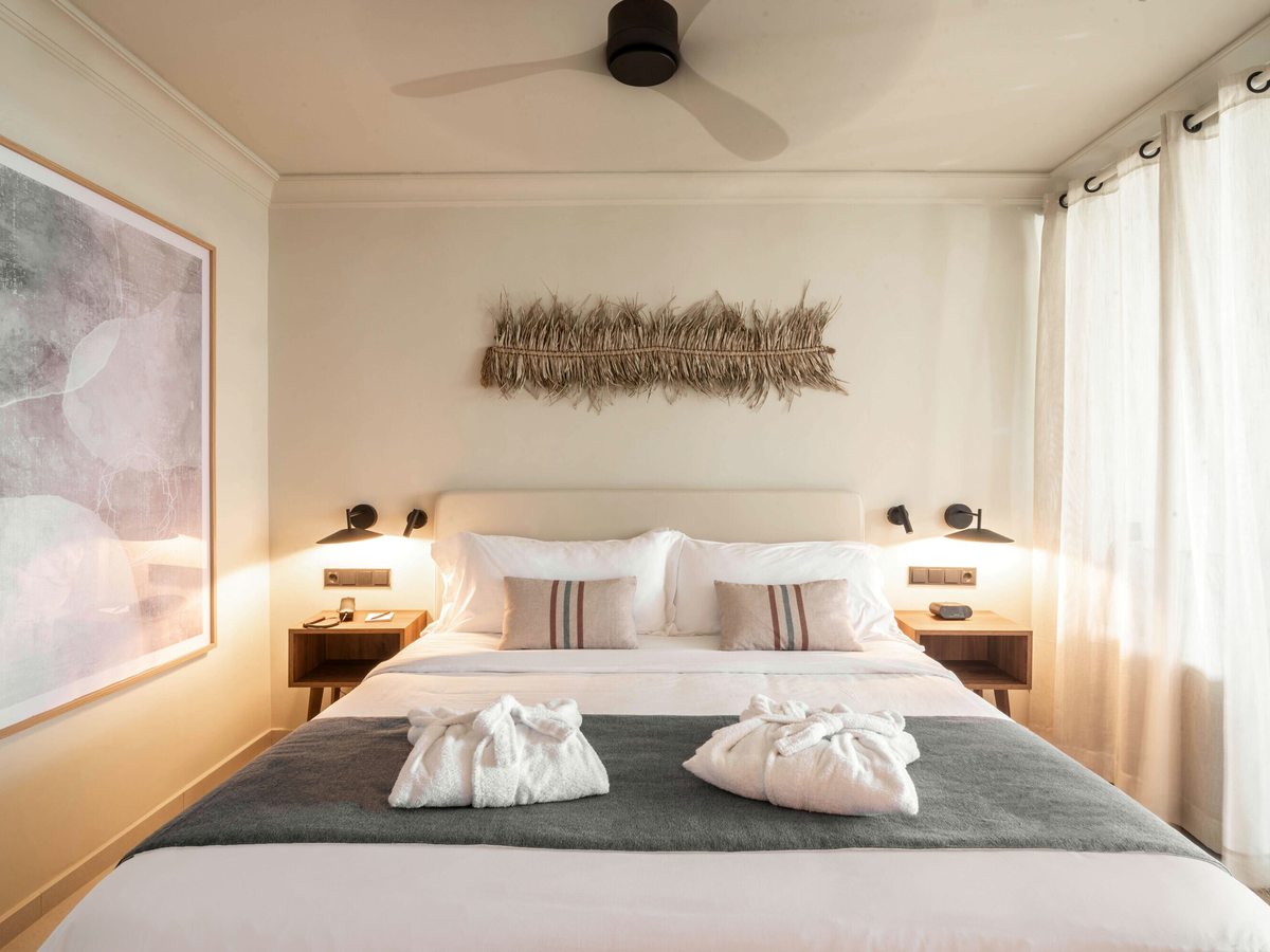 Double bedroom with a large bed, bathrobes laid out, reading lights, a ceiling fan and sheer curtains at large windows.