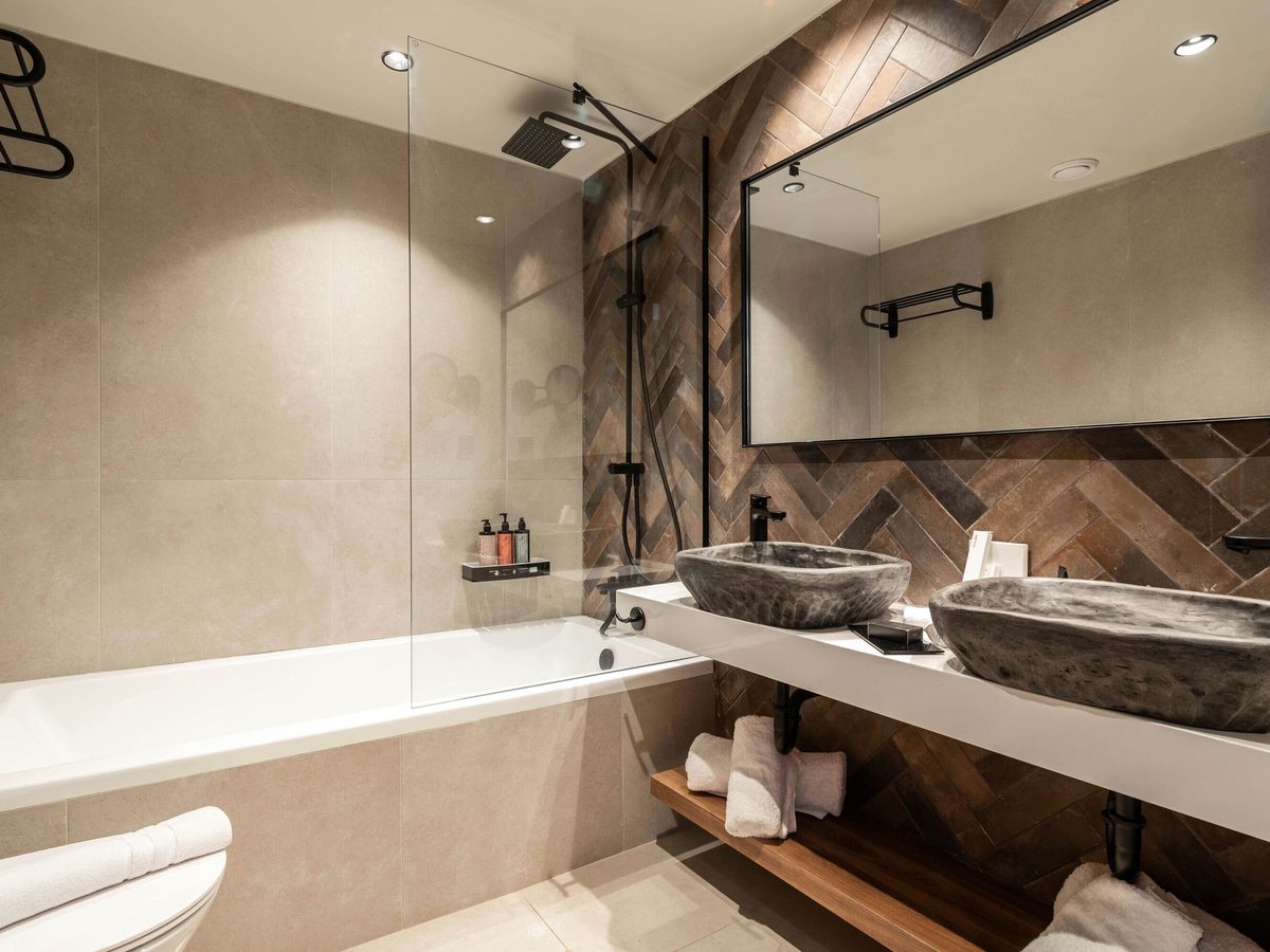 Bathroom with a double vanity, two stone vessel basins and a bathtub with a glass screen and shower over, on herringbone feature tiling.
