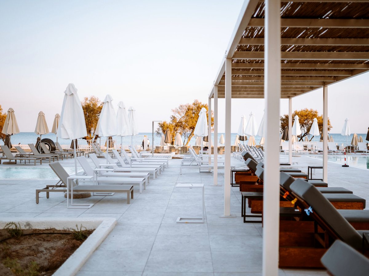 Sun loungers and white parasols along a seafront pool terrace beside a timber pergola, with planted trees and a sea view.