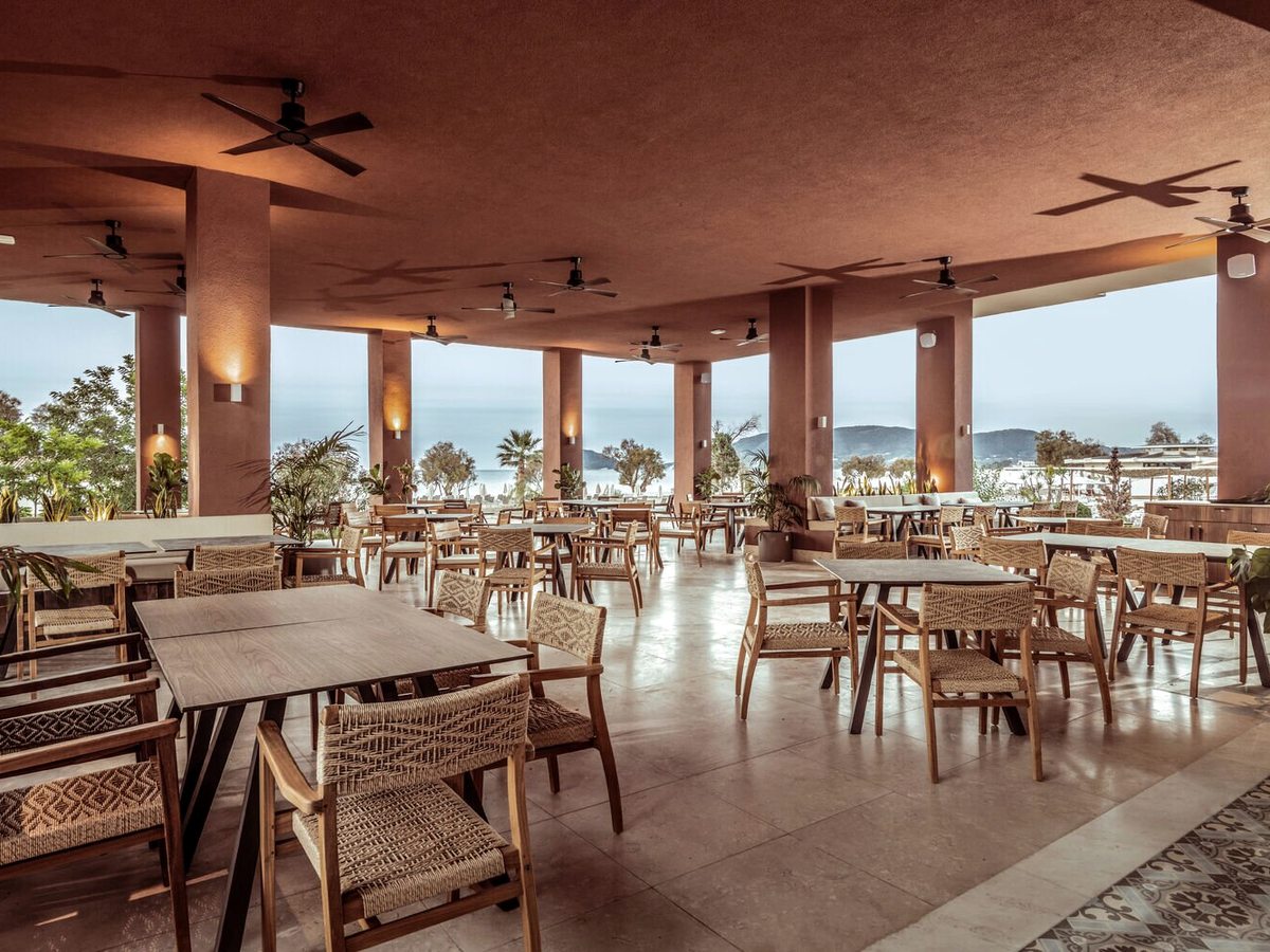 Covered open-air dining terrace with woven chairs and wooden tables under ceiling fans, looking out to palm trees, distant hills and the sea.