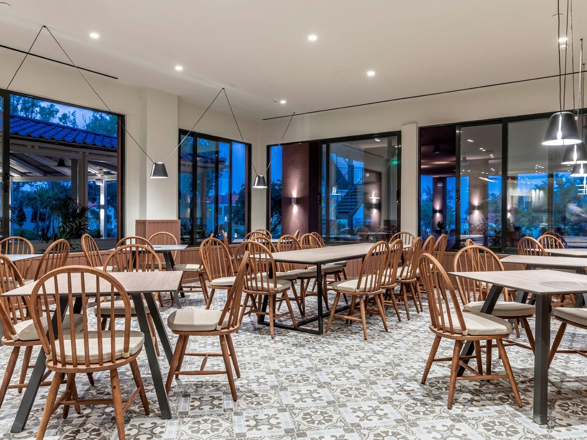 Indoor restaurant with wooden tables and spindle-back chairs on a patterned tiled floor under pendant lights, with floor-to-ceiling windows to a covered terrace.