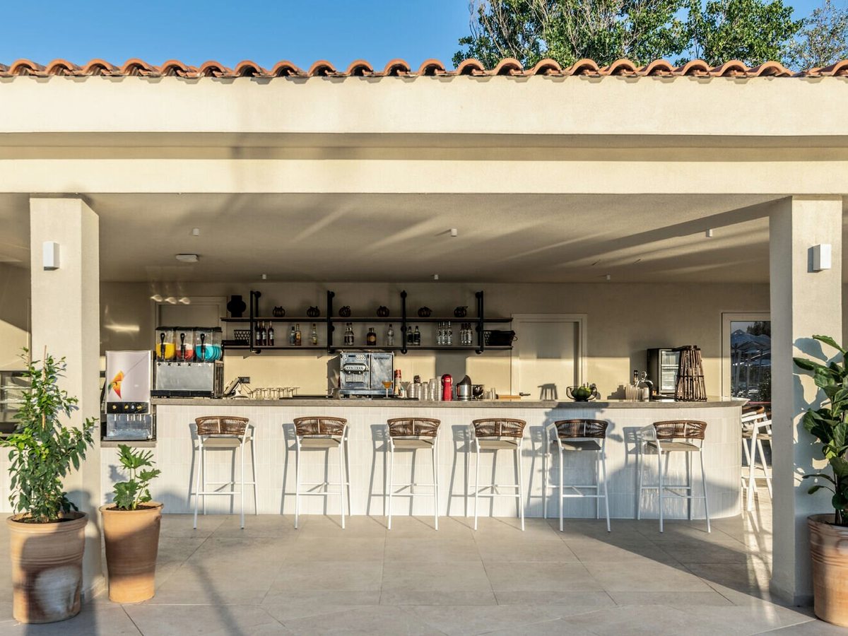 Open-air bar under a terracotta-tiled roof with a long counter, high stools, bottle shelves and potted tropical plants.