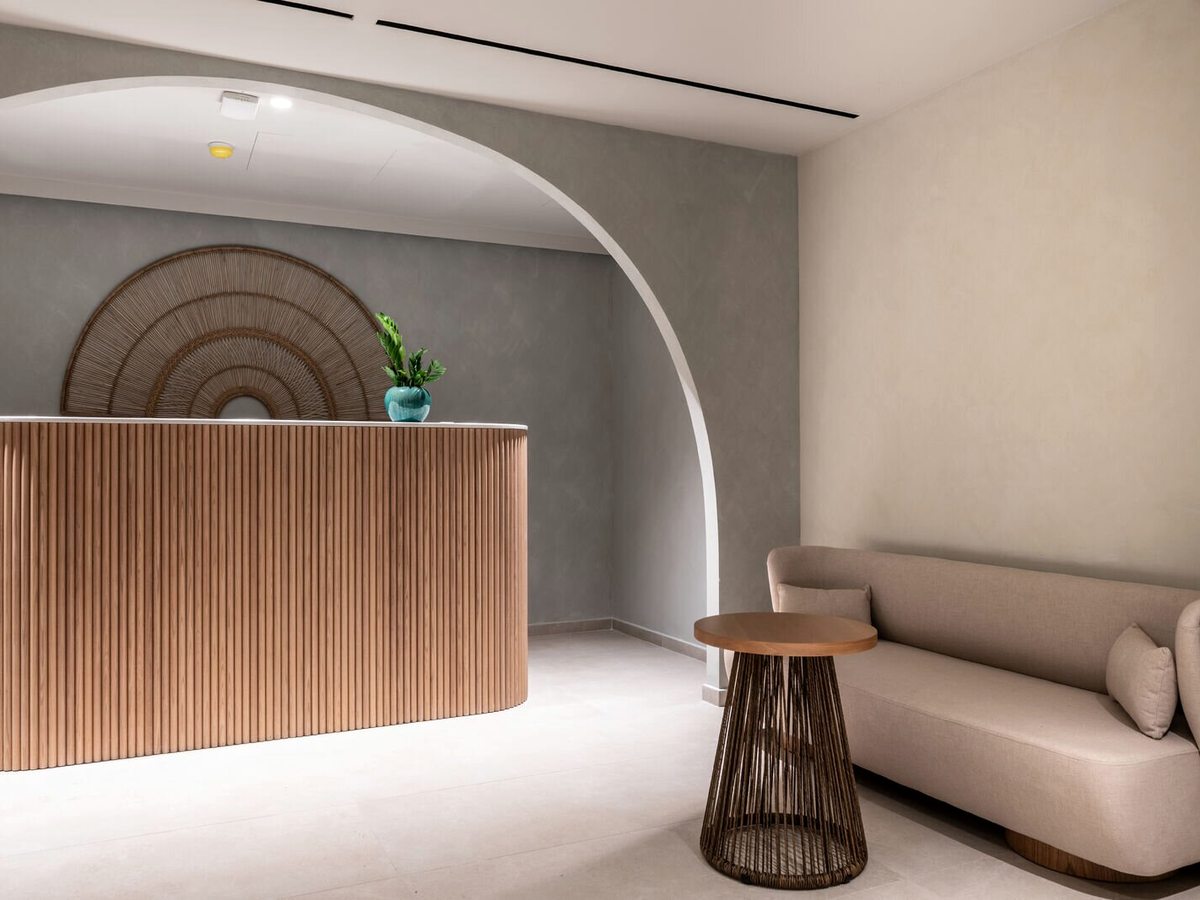 Curved timber reception desk with vertical wood slats under an illuminated arch, with a neutral sofa and woven side tables in a minimalist lobby.