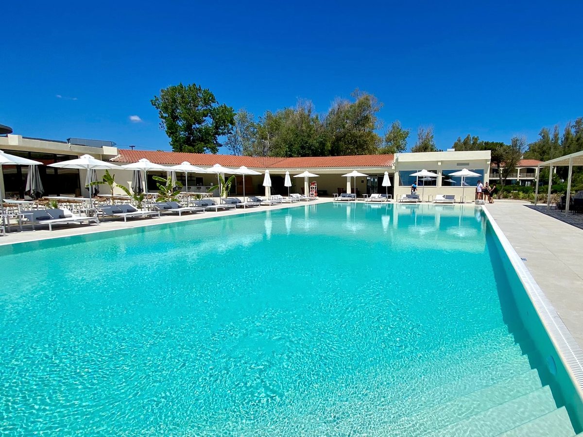 Domes Aulūs Zante's large turquoise outdoor pool with white parasols and sun loungers, low resort buildings behind.