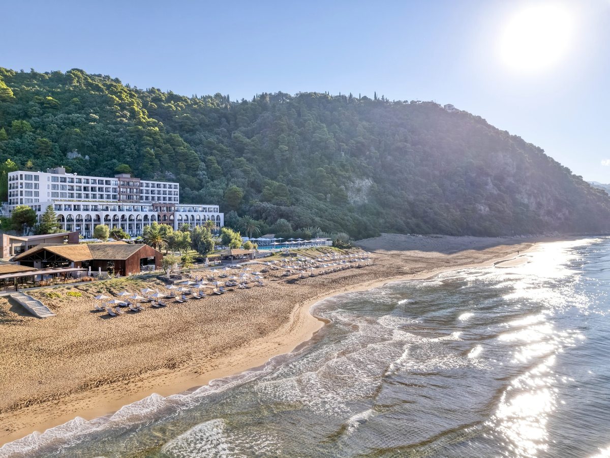Aerial view of the long sandy beach curving along the coast with the resort at one end, calm turquoise sea and a wooded headland.
