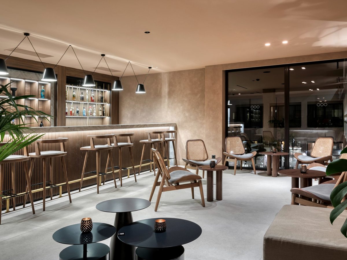 Indoor bar with a long timber counter and stools, cushioned armchairs and low tables under pendant lights, with an illuminated bottle display behind.