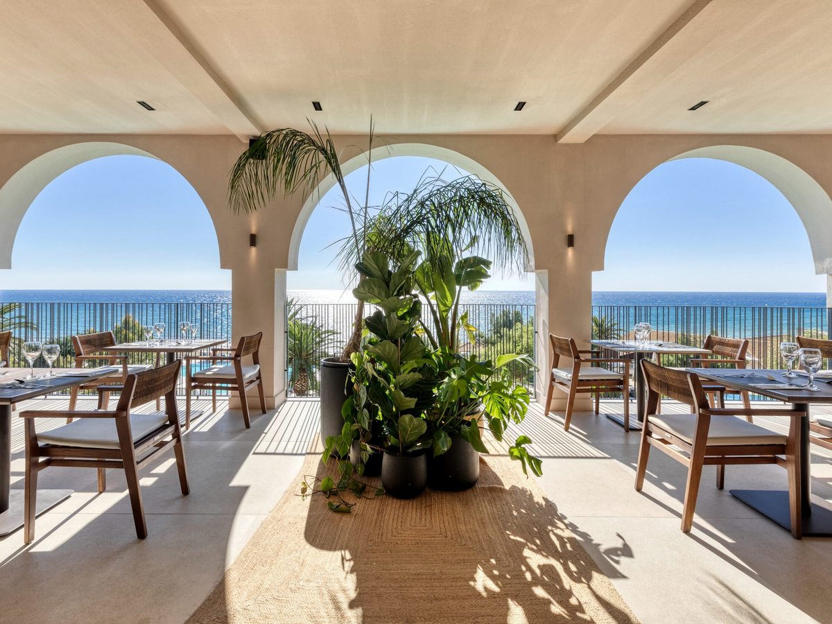 Covered dining terrace framed by three tall white arches, wooden tables and chairs set for a meal, tropical plants and an open sea view.