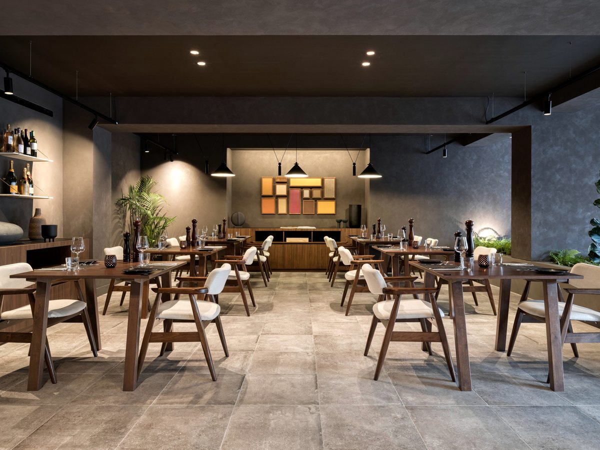 Contemporary indoor restaurant with widely spaced wooden tables and cream upholstered chairs under pendant lights, concrete-effect walls and a grey tiled floor.