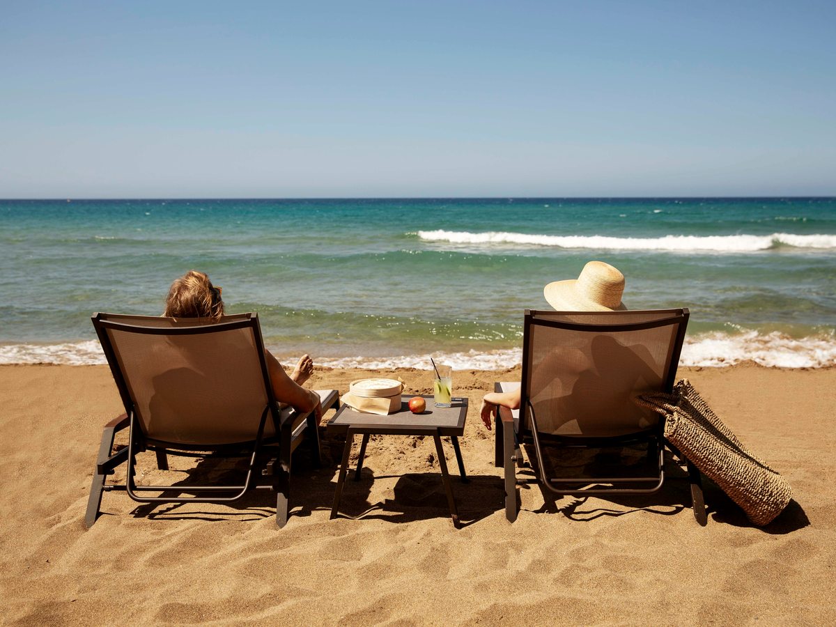 Cushioned beach loungers with a small table and a sun hat on the sand, looking out over the calm sea.