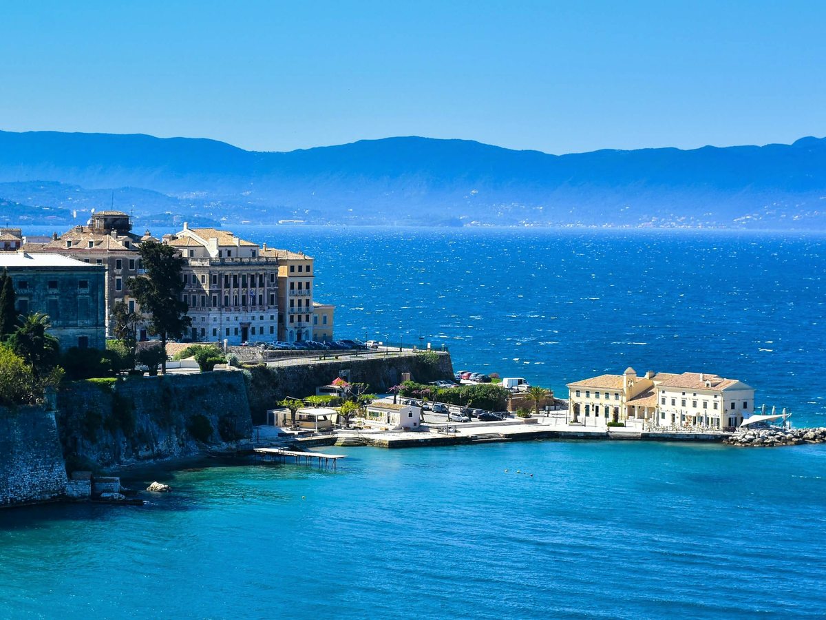 The waterfront of Corfu Old Town with historic stone buildings and a fortress on a tree-covered headland above a blue bay.