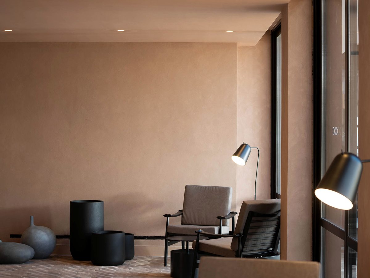 Neutral-toned indoor lounge with low armchairs, side tables and floor lamps beside floor-to-ceiling windows onto the garden.