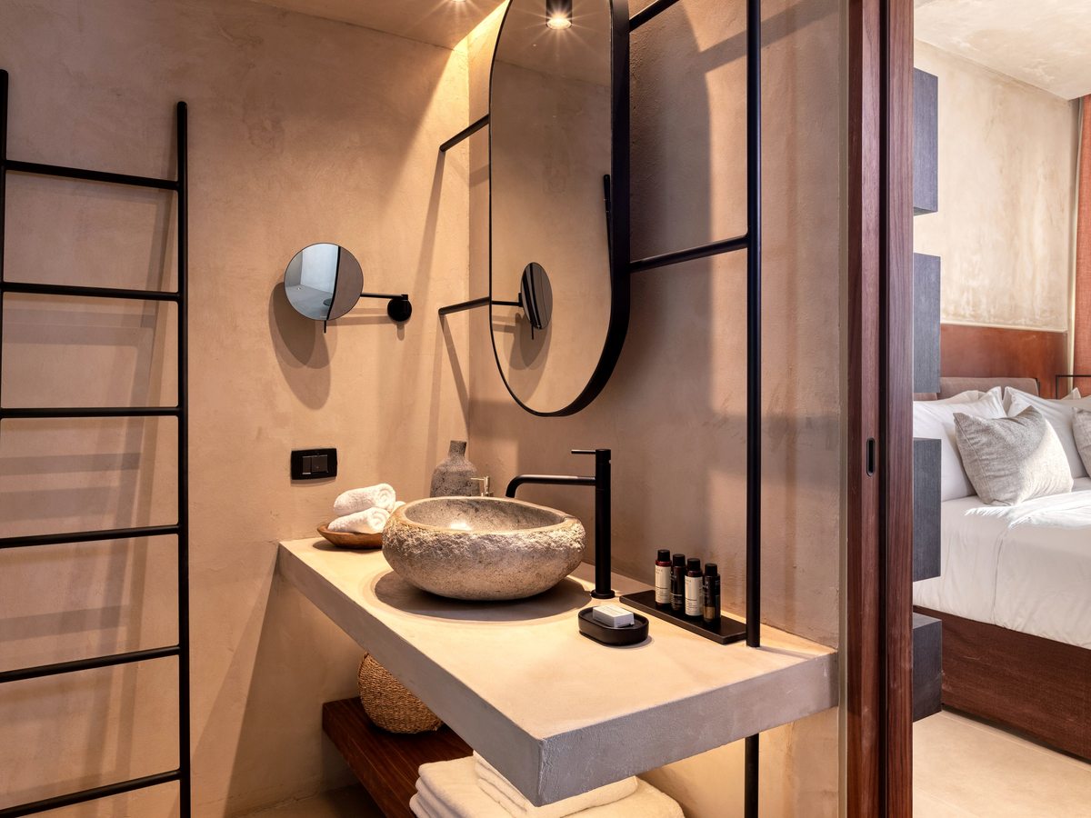 Open-plan bathroom with a stone vessel basin on an open vanity, a rounded mirror, a ladder towel rail and folded towels, beside the bedroom.