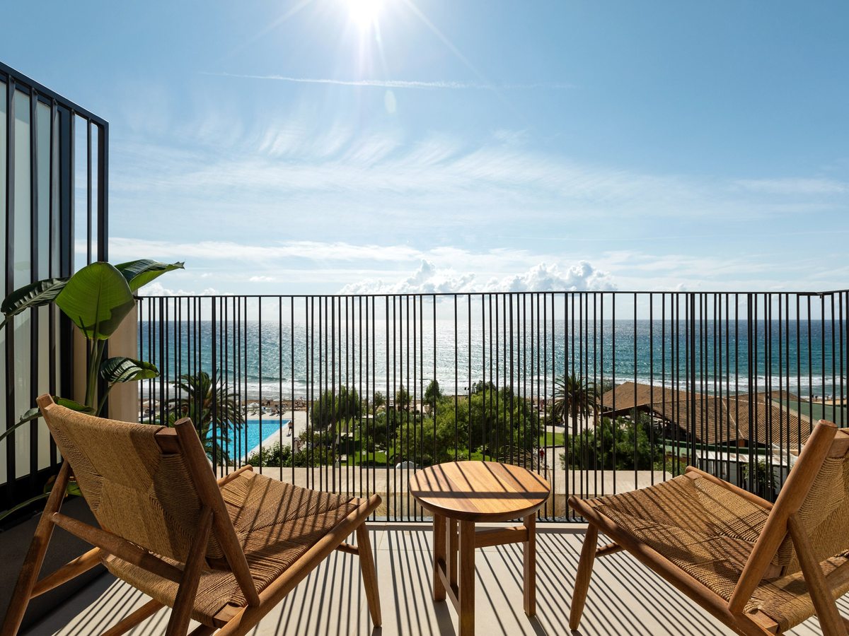 Elevated room terrace with woven lounge chairs and wooden side tables behind a metal railing, looking over the pool and the sea.