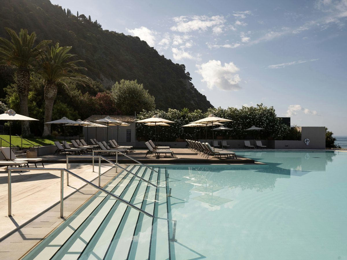 Broad submerged entry steps with stainless-steel handrails leading into a pool, sun loungers and parasols along the terrace and the sea beyond.
