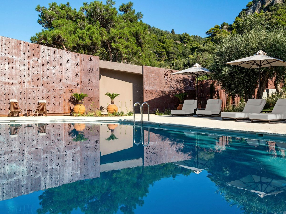 Rectangular pool enclosed by rust-coloured perforated screen walls, with cushioned sun loungers, parasols and side tables on a stone terrace among trees.