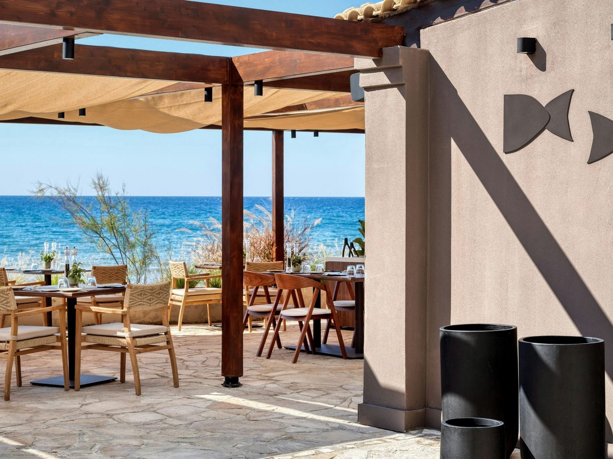 Open-air beachfront restaurant with wooden tables and woven chairs under a timber pergola and canvas shade sails, the sea beyond.