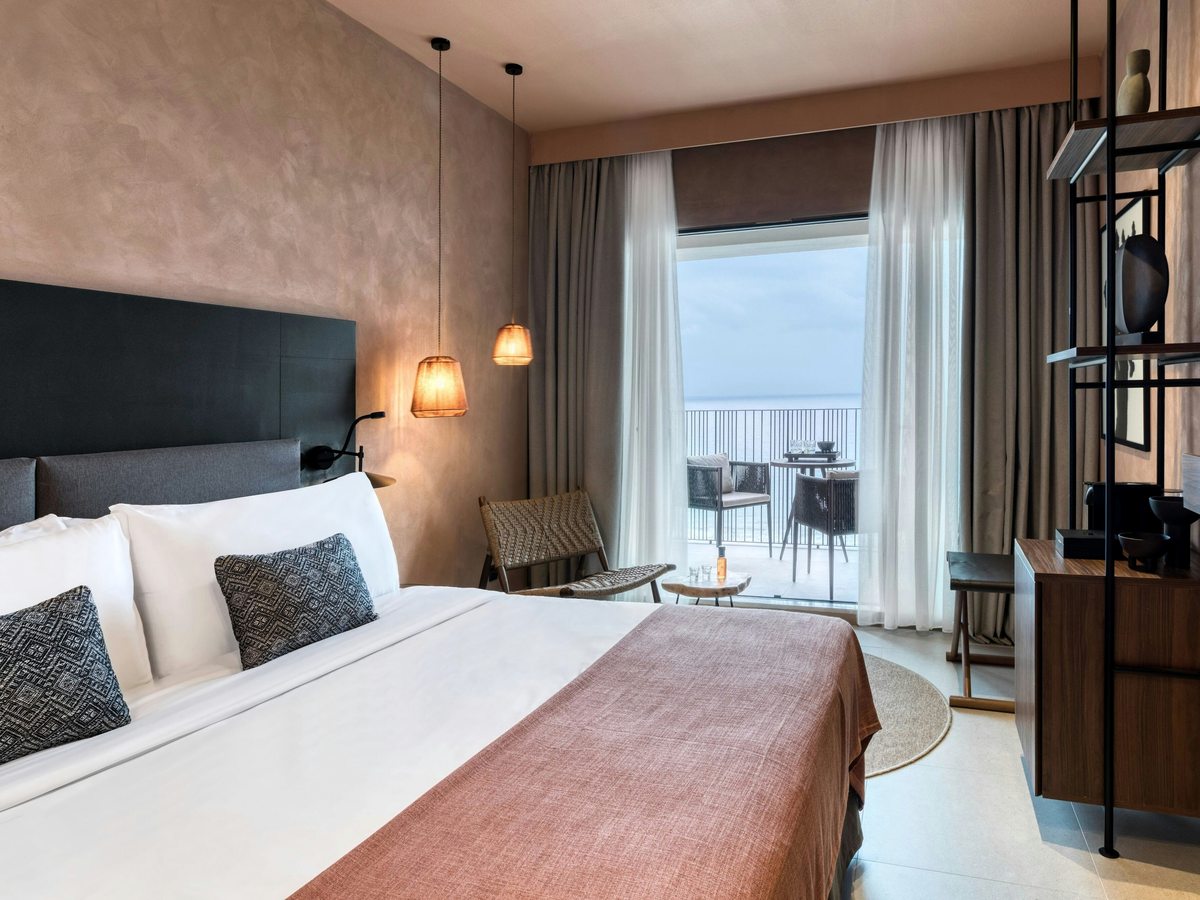 Double bedroom in warm neutral tones with a padded headboard, a wall-mounted television and open shelving, and glazed doors onto a furnished sea-view balcony.