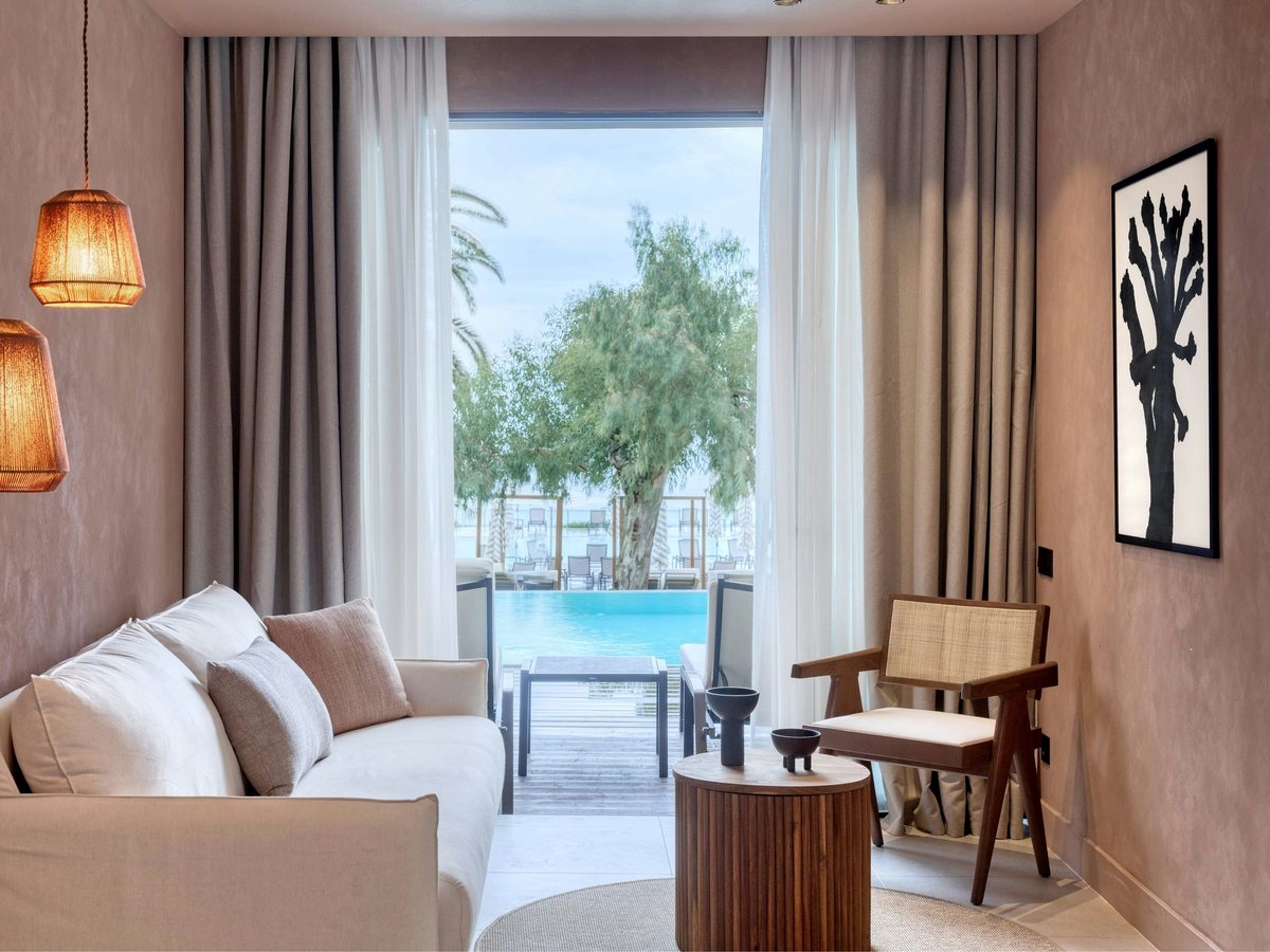 Ground-floor suite lounge with a white sofa and woven armchair opening through full-height glass doors onto a private terrace with direct access to a pool.