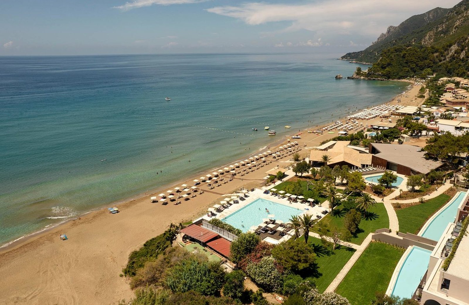 An aerial view of Domes of Corfu on the sandy Glyfada beach, its pools and gardens set below the wooded cliffs with the Ionian Sea beyond.