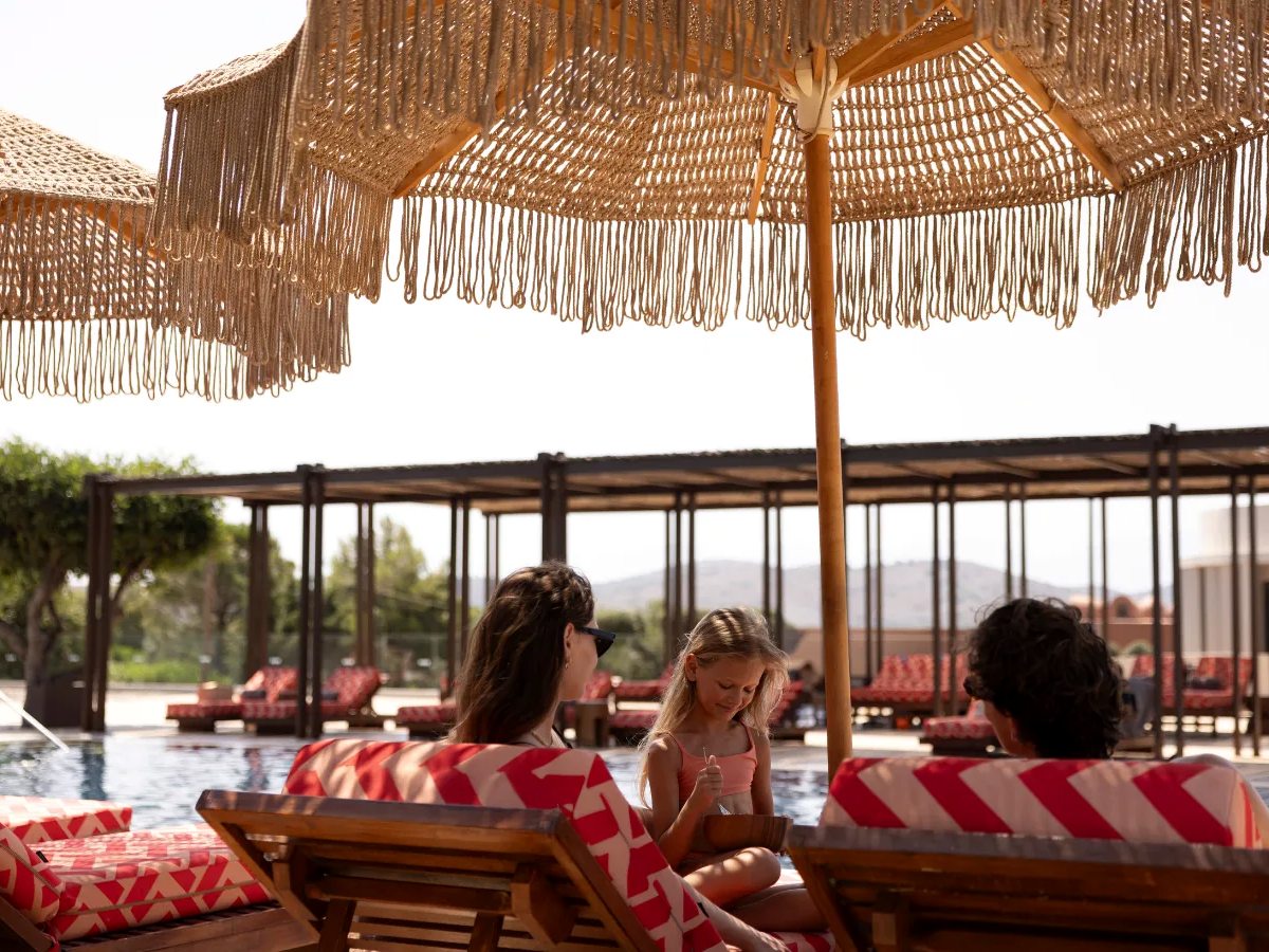 Wooden loungers with red-and-white cushions under a fringed straw parasol beside a pool, with cabanas and hills behind.