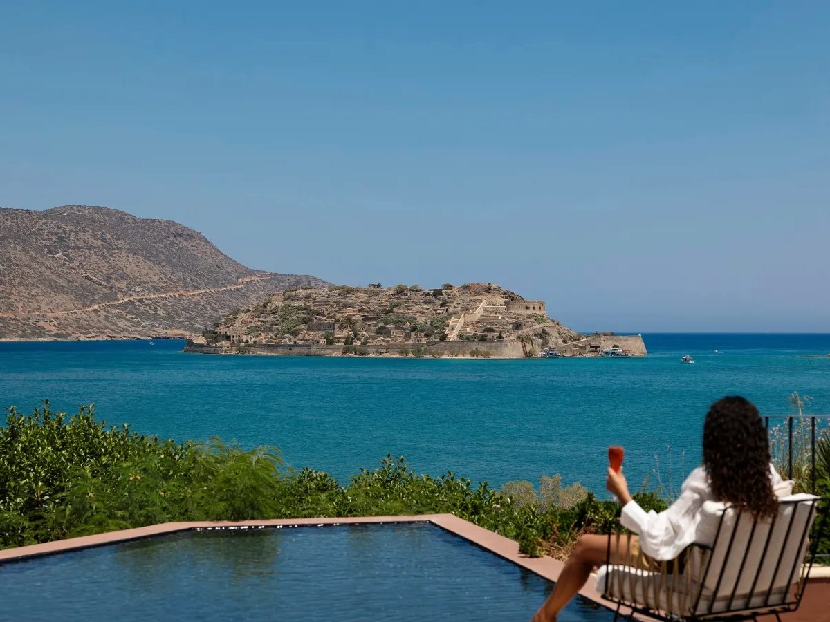 A dark infinity pool on a terrace with a single lounger, looking across the bay to the fortress on Spinalonga island.