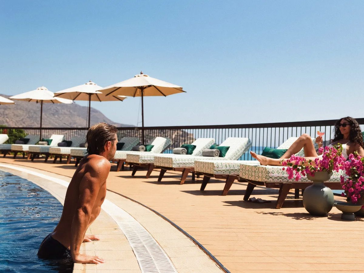 A curved sea-view pool edged with cream umbrellas and patterned loungers, with bougainvillea and the bay beyond.