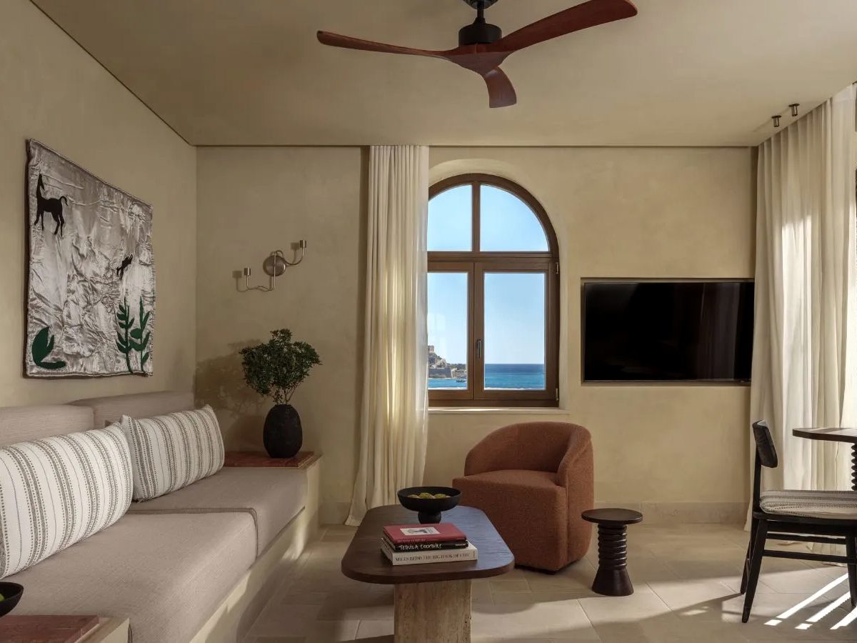 A suite living room with a pale sofa, a rust armchair and an arched window framing the sea, with a wall-mounted TV under a wooden ceiling fan.