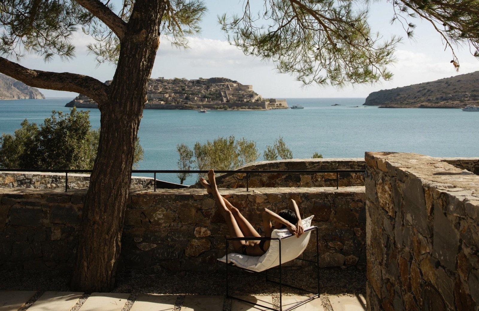 A stone terrace shaded by a pine tree at Domes of Elounda, looking across Mirabello Bay to the Venetian fortress on Spinalonga island.
