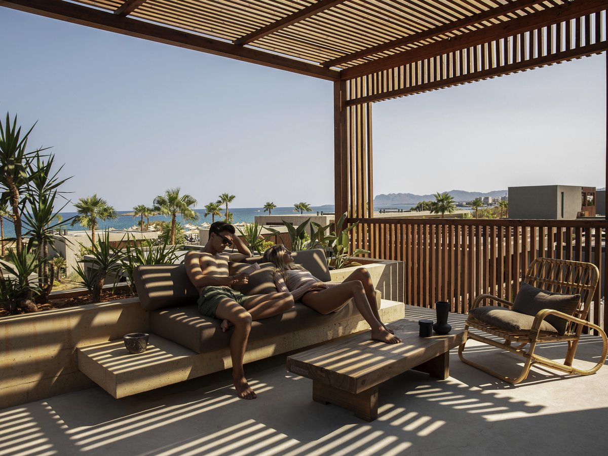 A timber-pergola terrace with a built-in daybed, looking out over the resort and palms to the sea.