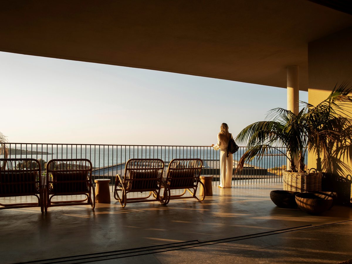 A stone terrace set with rattan armchairs under palms, opening to a long view down the hillside to the Aegean.