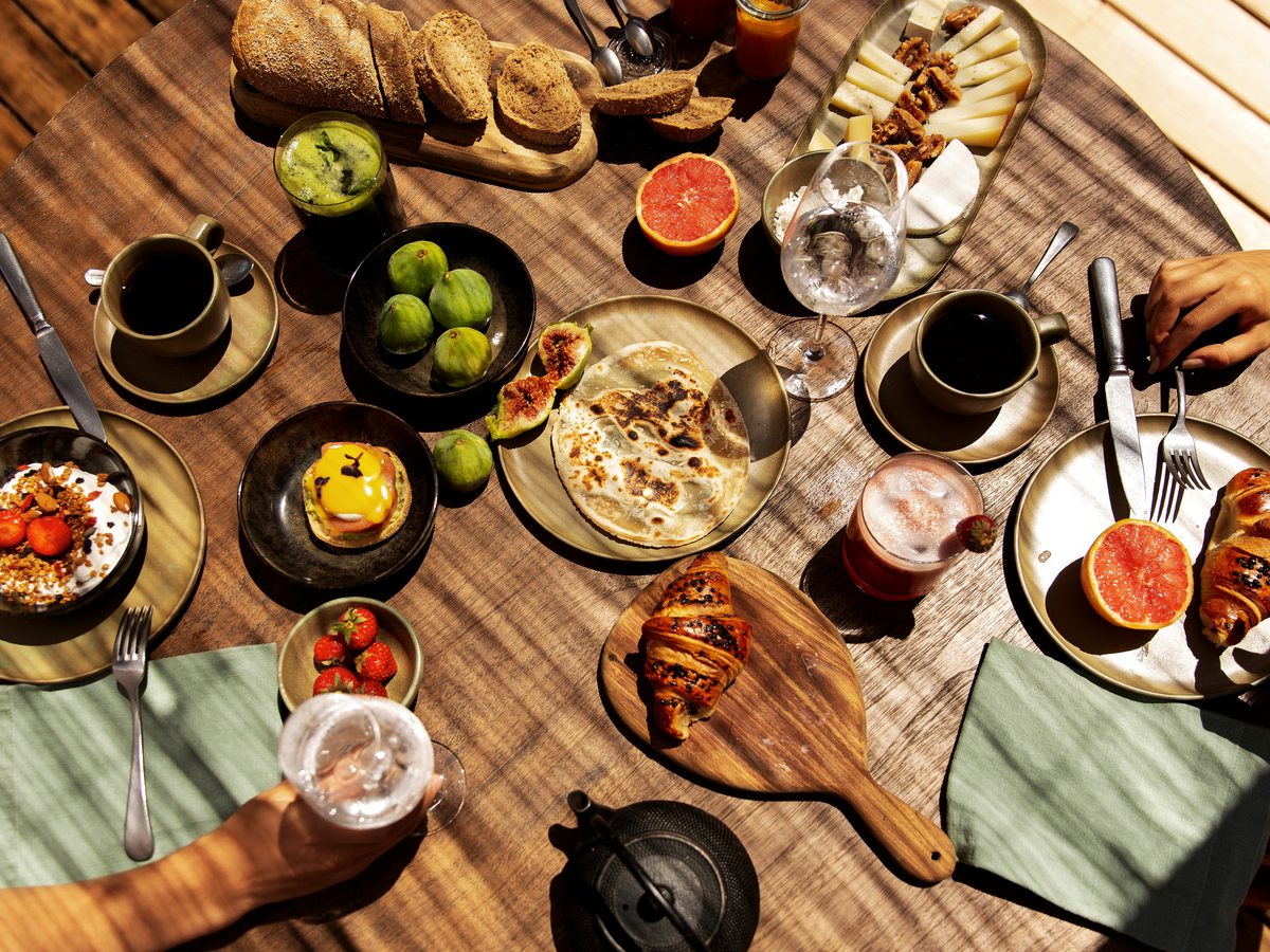 An overhead view of a breakfast spread of Greek dishes, breads, fruit and preserves laid across a wooden table.