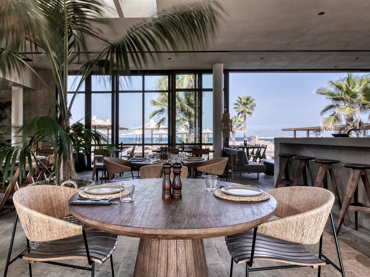 A restaurant with woven tub chairs around timber tables, opening through full-height glass to palms, thatched umbrellas and the sea.