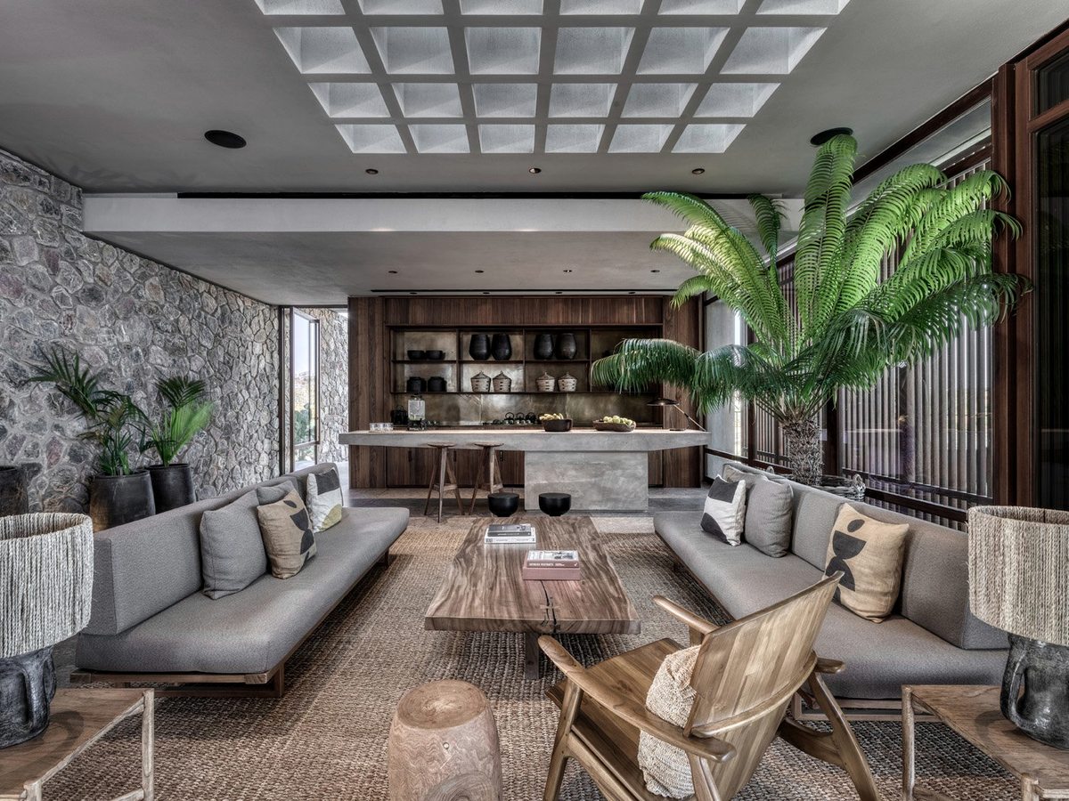 An open-plan indoor lounge with low grey sofas, a timber ceiling and a stone feature wall, potted palms and a sea view beyond.