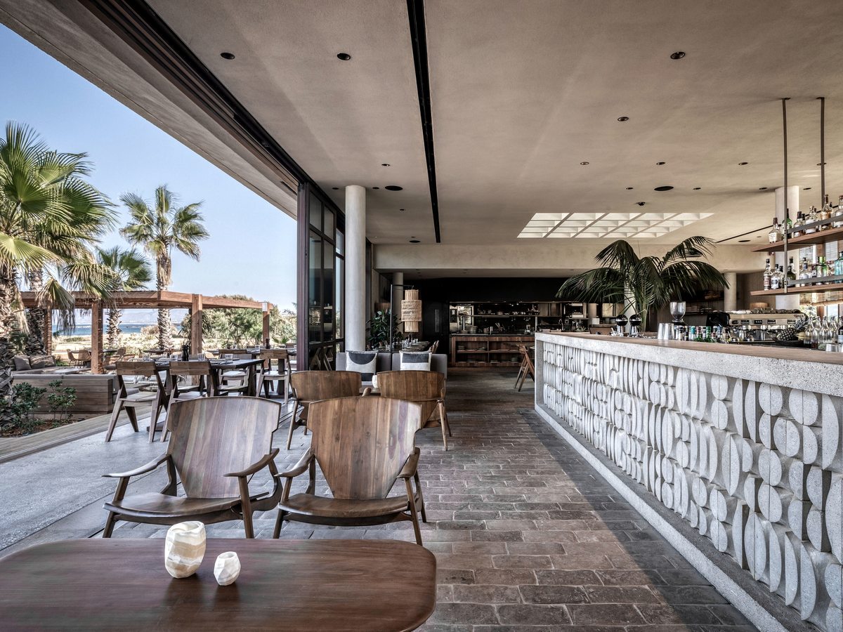 A long bar with a carved pale-stone front and back-lit spirits shelves, opening to a terrace of palms and sea.