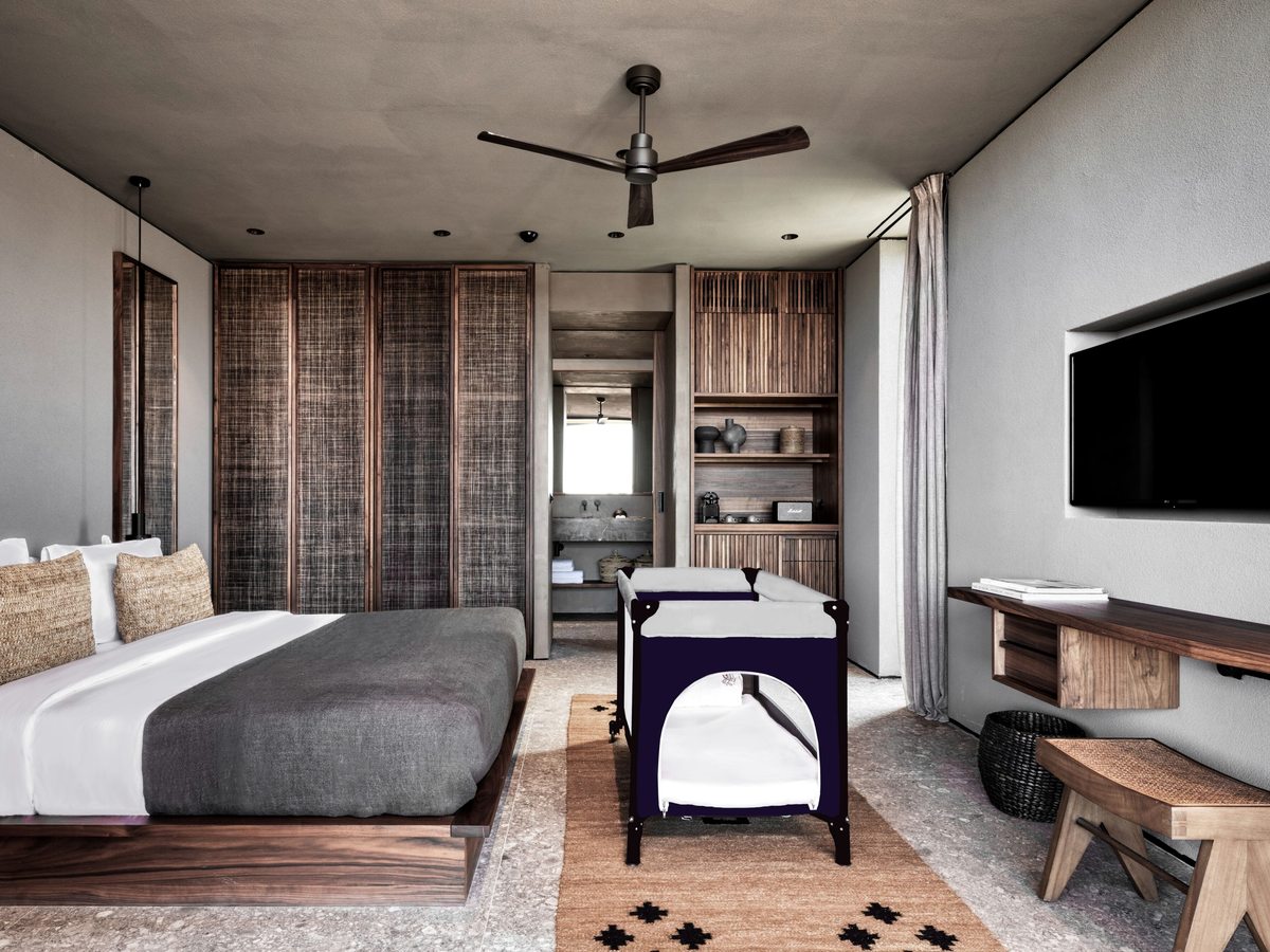 A bedroom with a king bed and a travel cot at its foot, a slatted timber wardrobe wall and an open bathroom beyond.