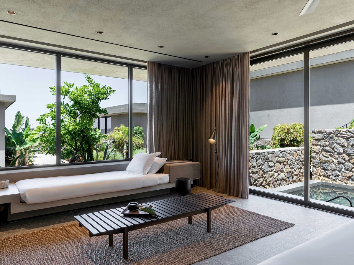 A bedroom with floor-to-ceiling glass on two sides, opening to a small private plunge pool set against a stone wall and greenery.