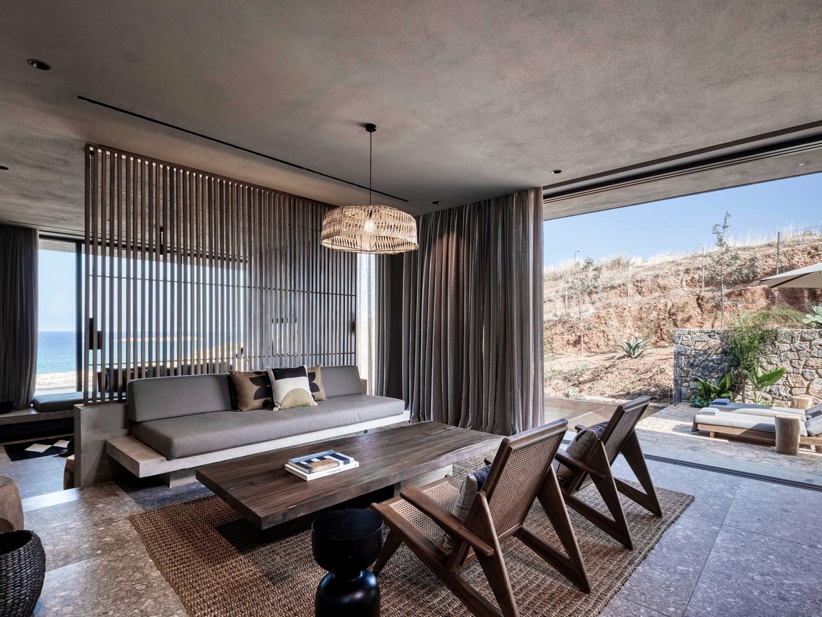 A suite living room with a grey sofa and cane chairs, floor-to-ceiling glass to the sea on one side and a walled terrace with loungers on the other.