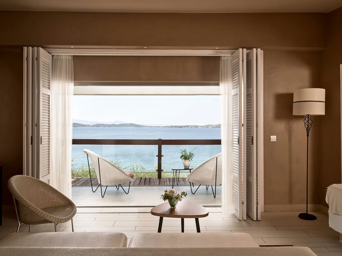 Room sitting area with a wicker chair and a low coffee table, folding shutter doors open to a sea-view terrace with two chairs.
